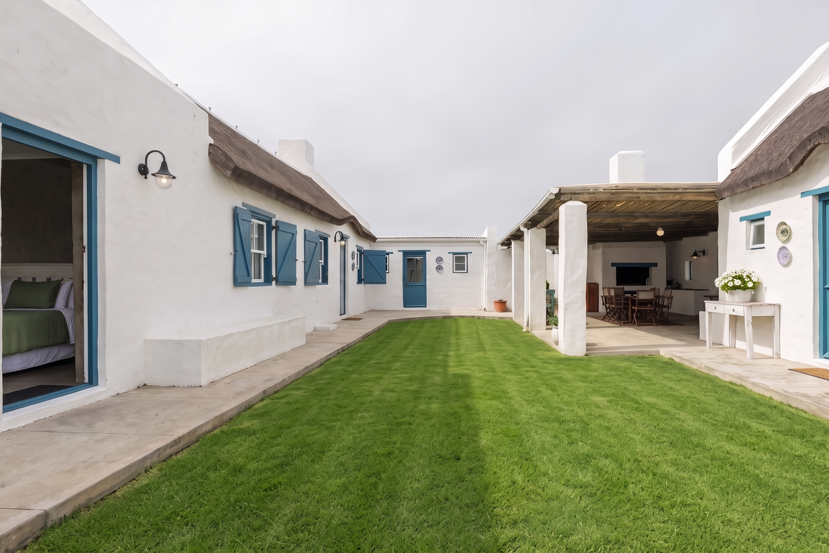 A well-maintained rear courtyard featuring a manicured lawn, a covered patio with a built-in fireplace, and charming Cape Dutch style architecture with thatched roofing.