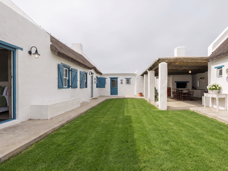 A well-maintained rear courtyard featuring a manicured lawn, a covered patio with a built-in fireplace, and charming Cape Dutch style architecture with thatched roofing.