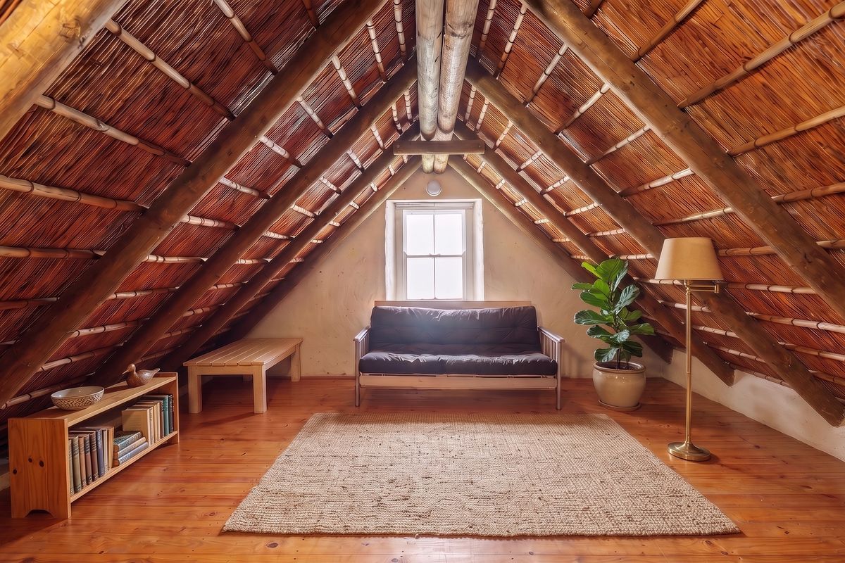 Attic bedroom featuring exposed timber rafters, a traditional thatched roof, polished timber flooring, and a central sash window.