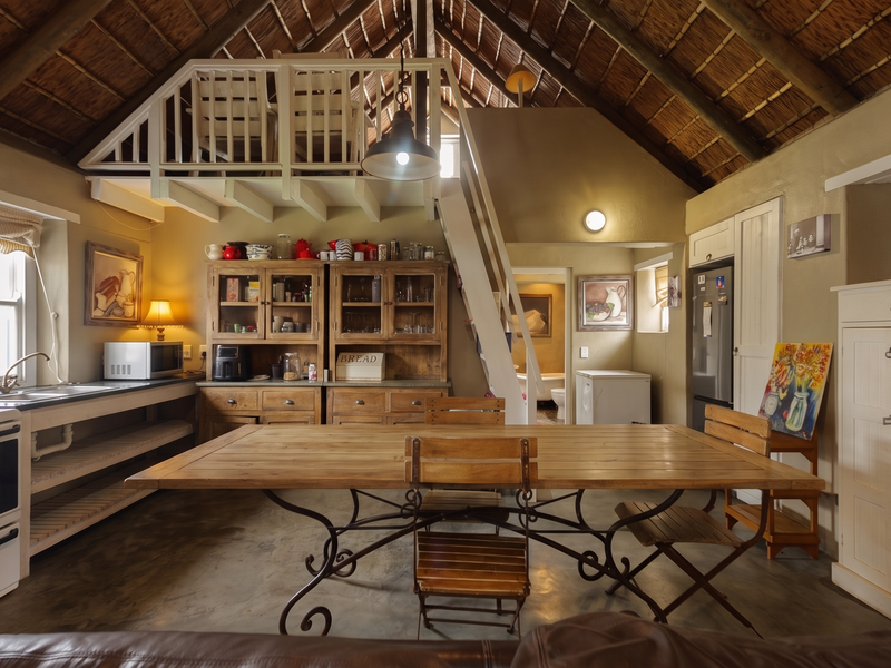 Open-plan kitchen and dining area featuring a rustic thatched roof, exposed timber rafters, a mezzanine level, and durable concrete flooring.