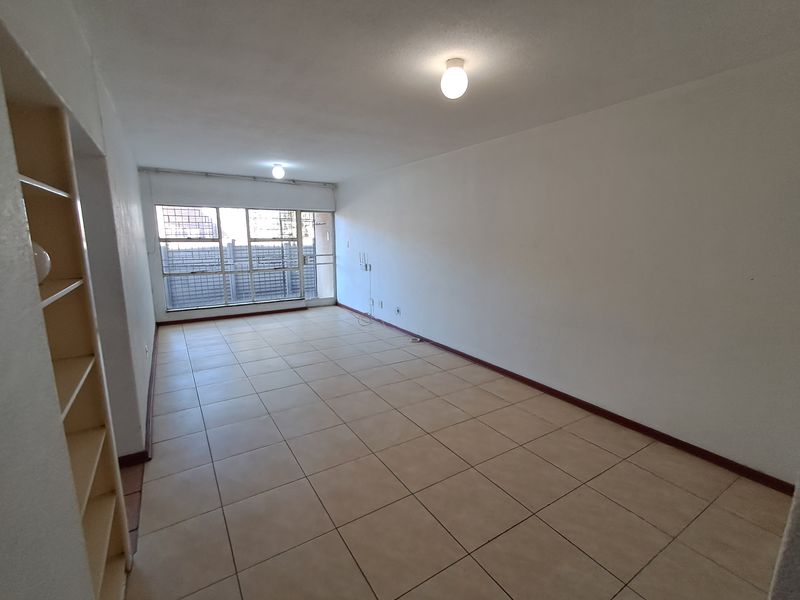 A spacious, empty living area featuring neutral tiled flooring, built-in shelving, and large windows that provide ample natural light.