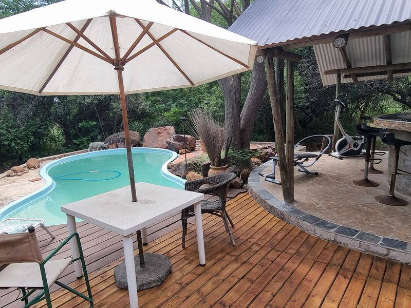 Outdoor entertainment area featuring a swimming pool, wooden decking, and a covered patio with a built-in bar, set in a natural bushveld environment.