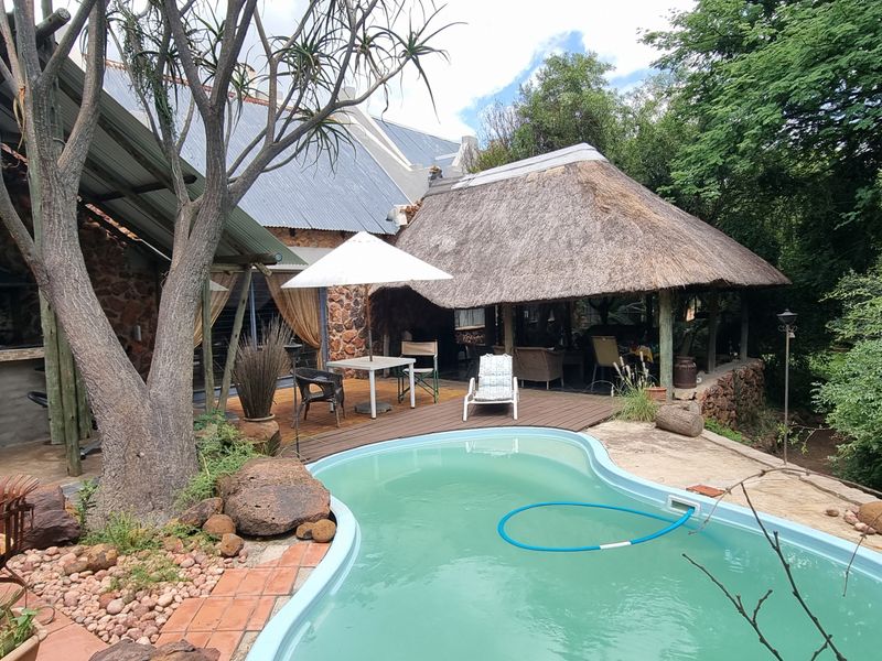 Outdoor entertainment area featuring a swimming pool, timber decking, and a rustic thatched lapa with a built-in braai, set within a natural garden.