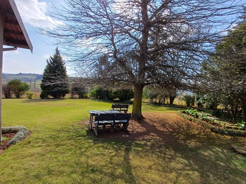 A spacious, well-maintained garden featuring a large mature tree, manicured lawn, and established garden beds with scenic rural views.