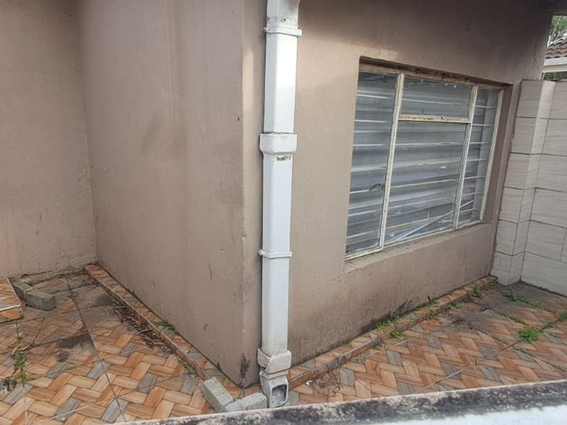 Rear exterior area of the cottage featuring a tiled patio, aluminium window, and boundary wall, showing signs of wear and maintenance requirements.