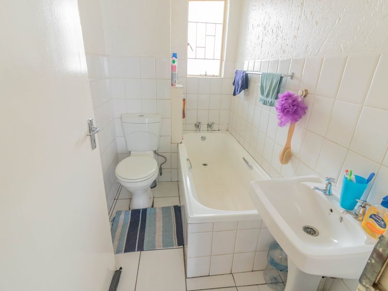 A compact bathroom featuring a standard bathtub, pedestal basin, and toilet with white tiled finishes and a window with security bars.
