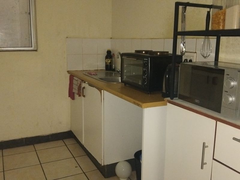 A compact kitchen area featuring basic cabinetry, a stainless steel sink, tiled splashback, and ceramic floor tiles, requiring cosmetic updates.