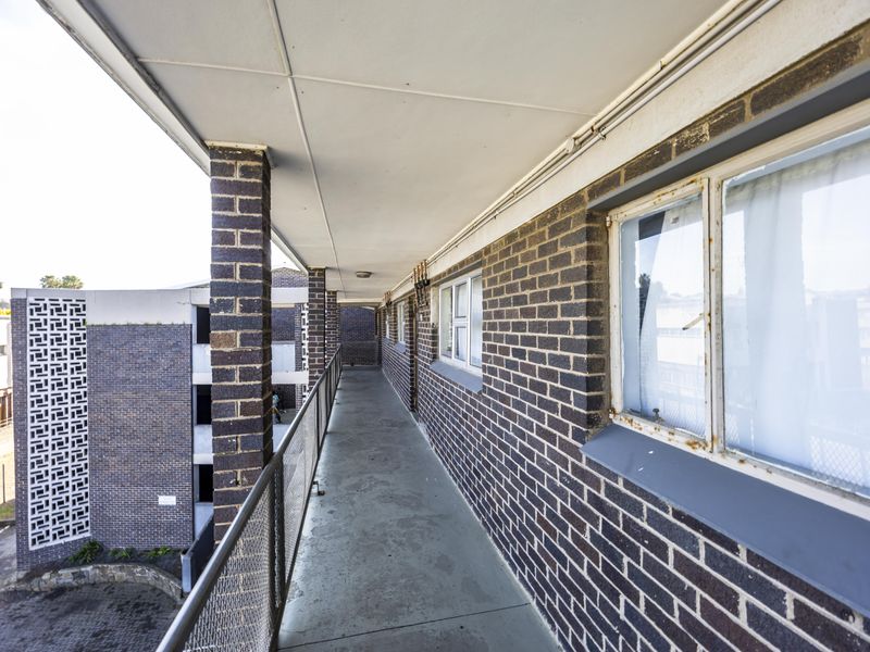 An exterior balcony walkway featuring brick walls, metal balustrades, and concrete flooring, showing signs of age and wear on the window frames.
