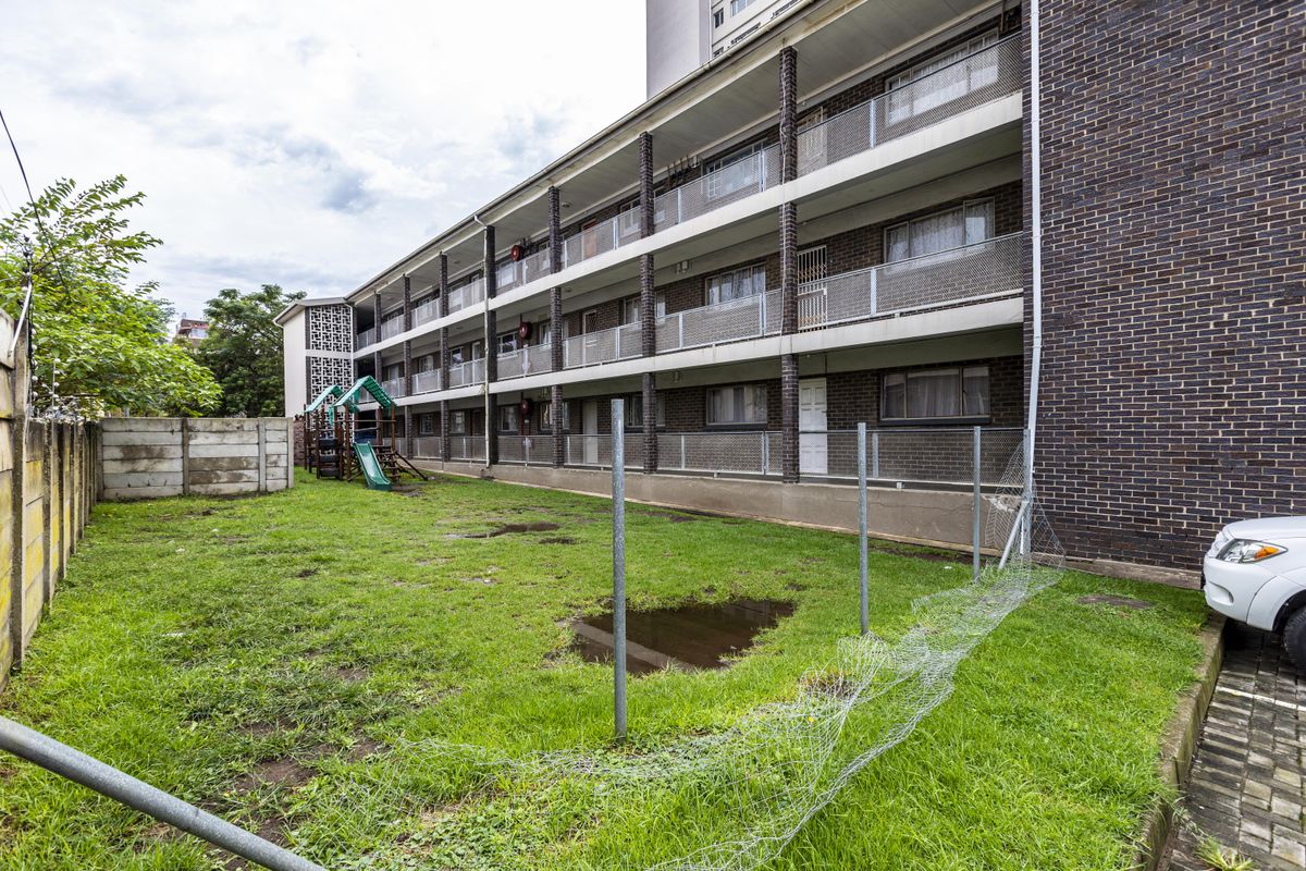 Rear view of a multi-story apartment building featuring brick exterior, tiered balconies with metal railings, and a shared lawn area with perimeter fencing.