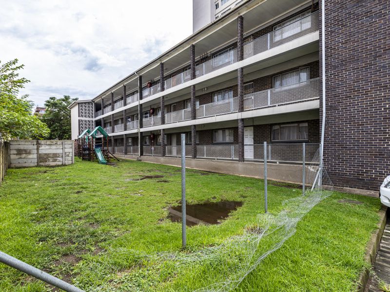 Rear view of a multi-story apartment building featuring brick exterior, tiered balconies with metal railings, and a shared lawn area with perimeter fencing.