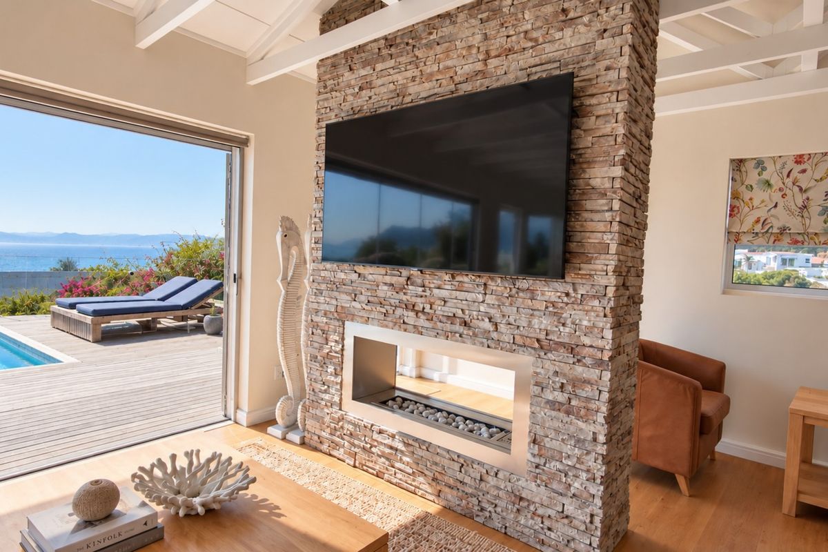 Bright living room featuring a striking stone-clad fireplace, exposed beam ceilings, and large sliding doors opening to a pool deck with ocean views.