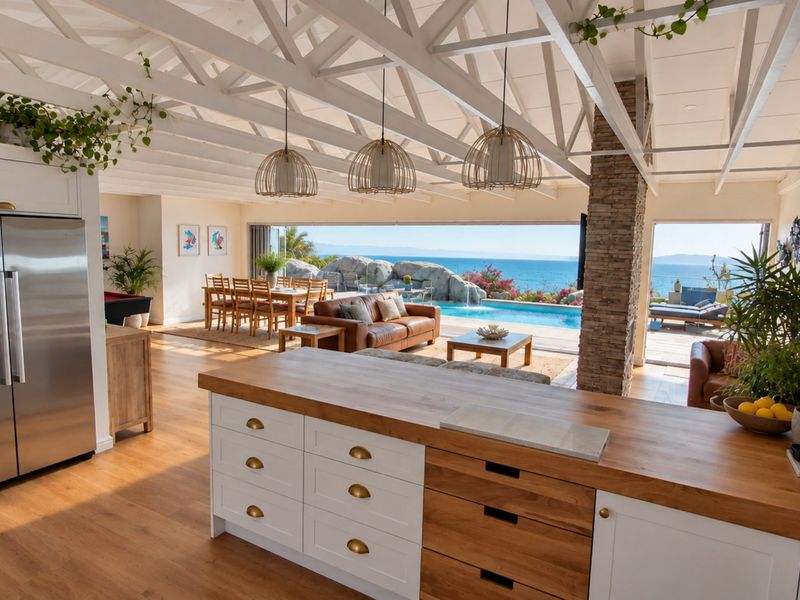 Spacious open-plan living area featuring a modern kitchen with island, exposed roof trusses, and seamless flow to the outdoor pool deck and ocean view.