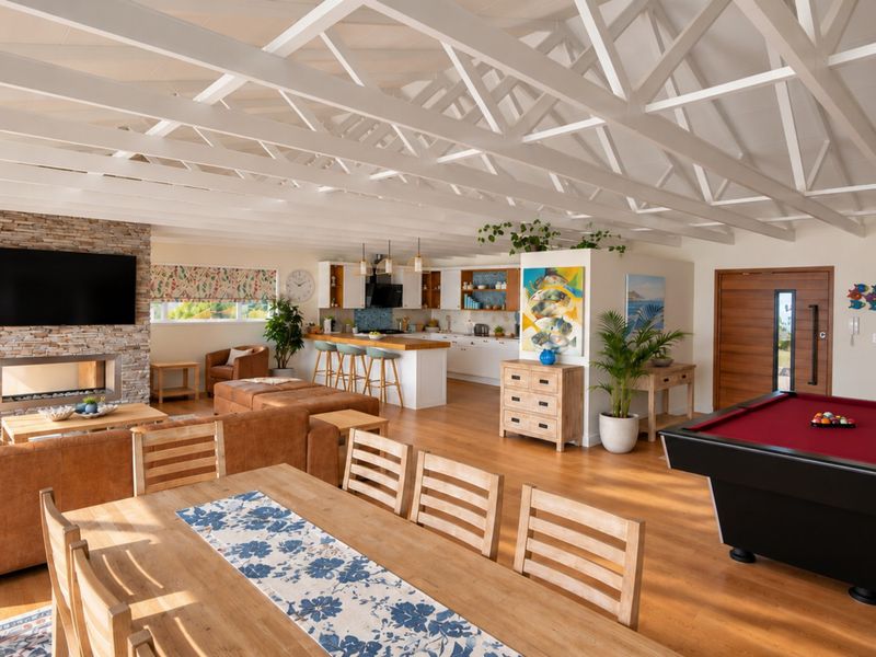 Spacious open-plan living area featuring a high exposed truss ceiling, stone fireplace, and a modern kitchen with a central island and hardwood flooring.