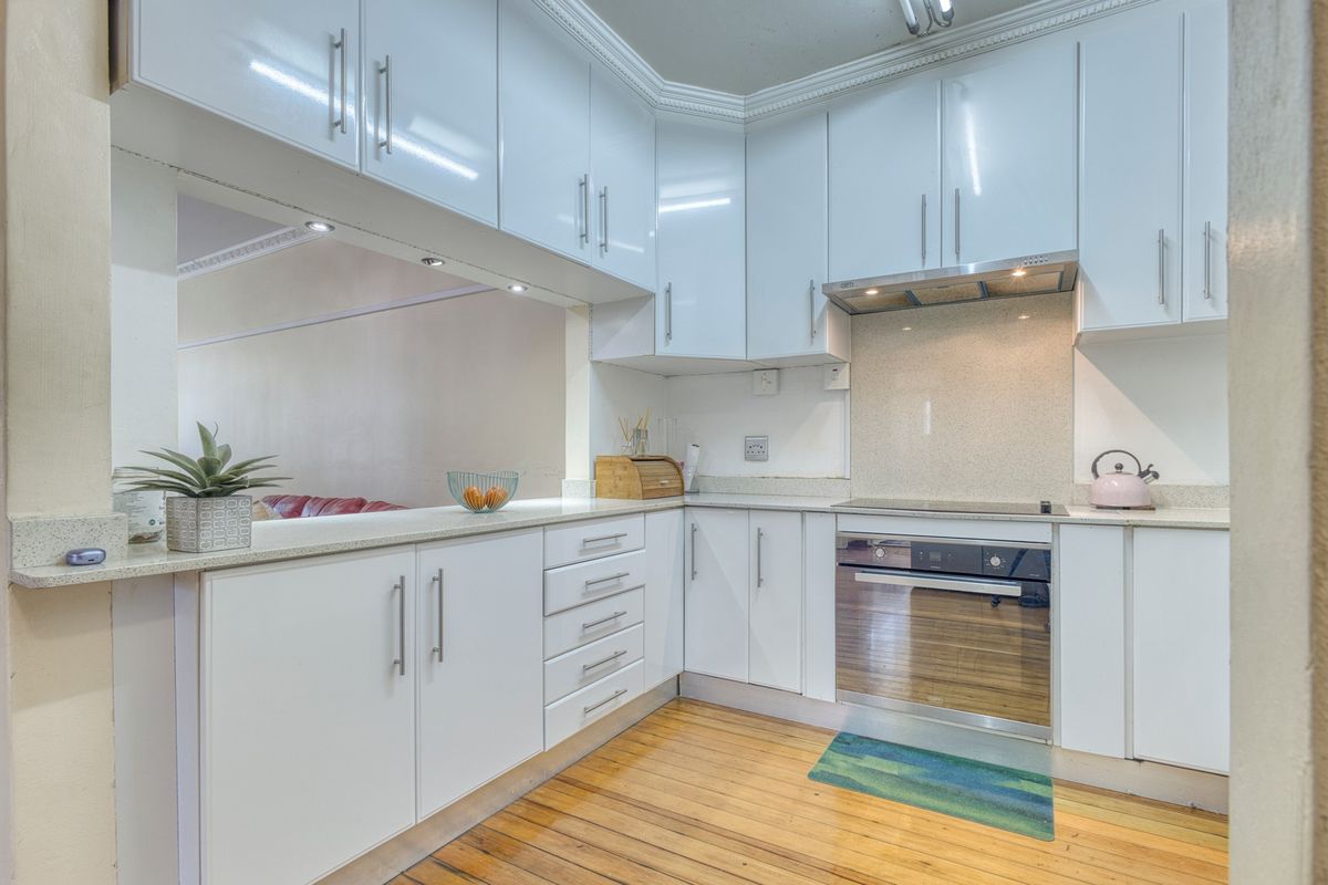 A modern, well-maintained kitchen featuring white gloss cabinetry, stone countertops, integrated oven and hob, and warm timber flooring with a convenient breakfast bar.