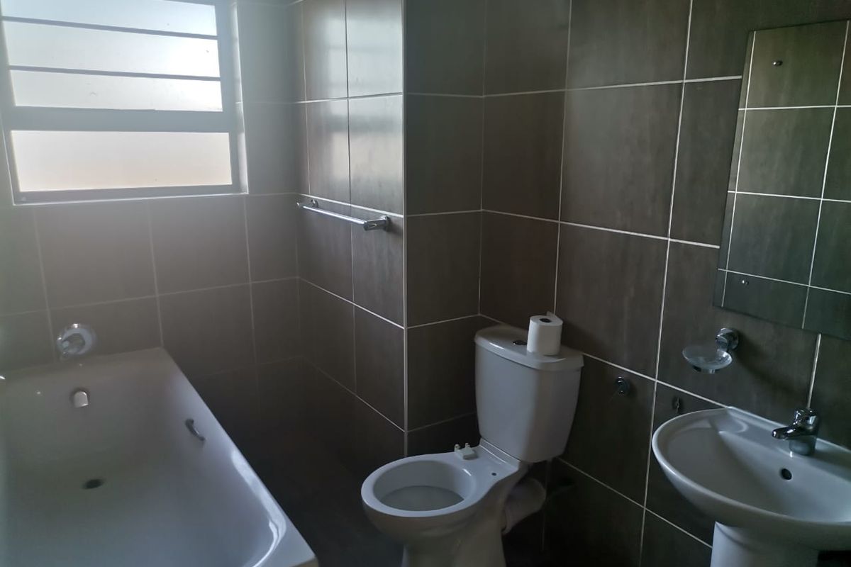 A functional bathroom featuring a bathtub, pedestal basin, and toilet with dark wall tiling and natural light from a frosted window.
