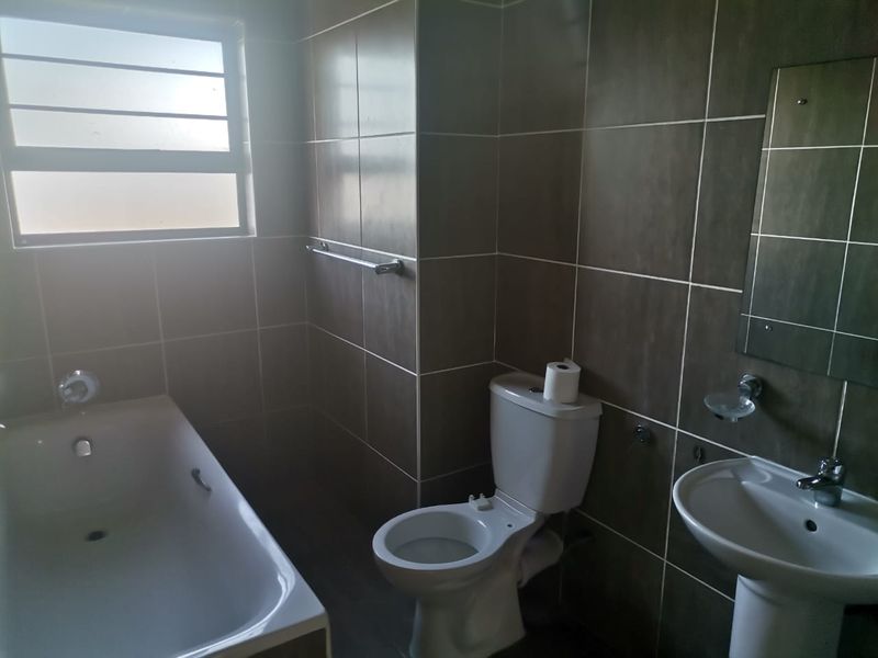 A functional bathroom featuring a bathtub, pedestal basin, and toilet with dark wall tiling and natural light from a frosted window.