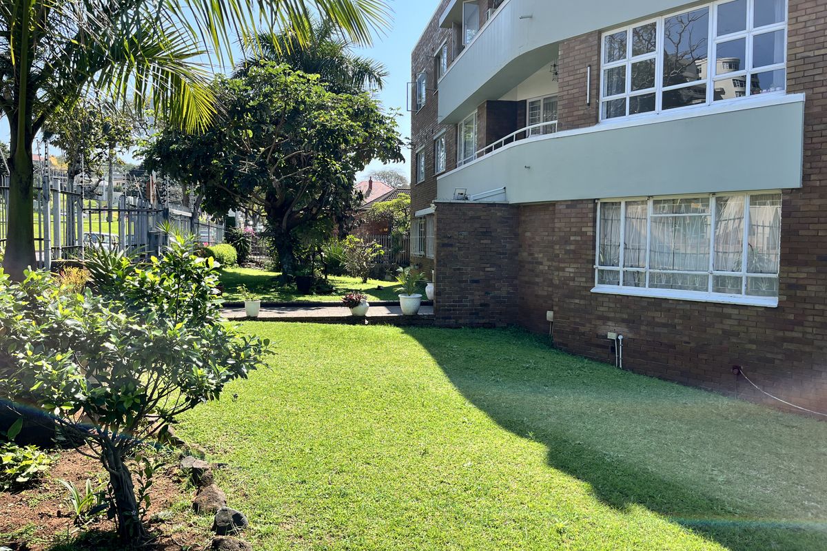 Rear view of a brick apartment building featuring a well-maintained garden, paved pathways, and balconies with metal railings.