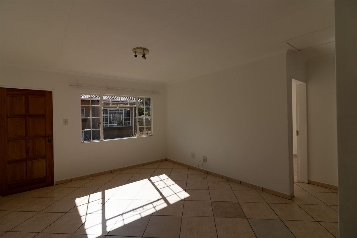 An empty, sunlit living room featuring neutral tiled flooring, a timber door, and a window with security bars.