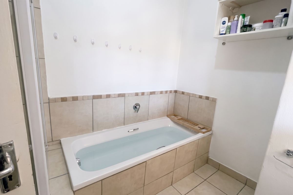 A functional bathroom featuring a built-in bathtub with tiled surround, wall-mounted shelving, and neutral floor tiling.