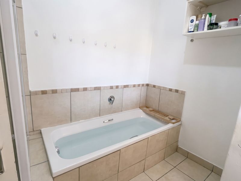 A functional bathroom featuring a built-in bathtub with tiled surround, wall-mounted shelving, and neutral floor tiling.