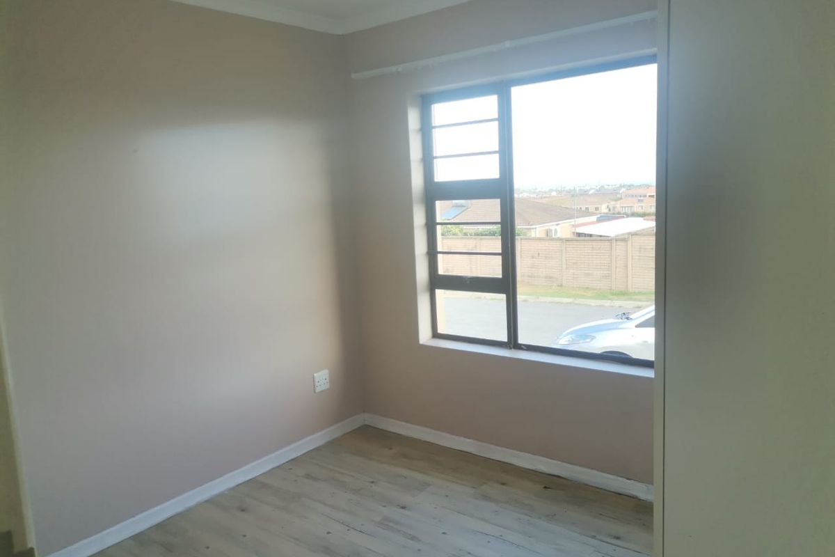 A bright, empty bedroom featuring laminate flooring, neutral wall tones, and a large window with security bars overlooking the street.