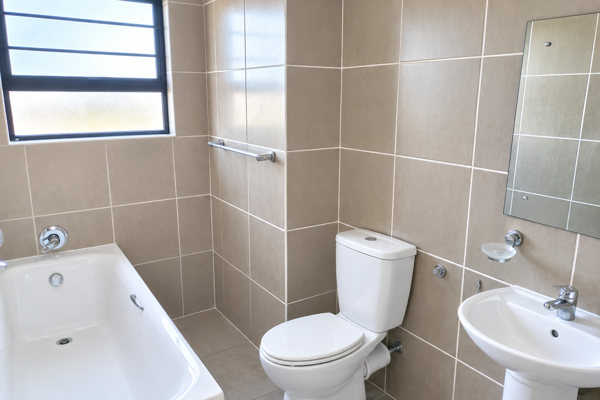 A clean, neutral-toned bathroom featuring a bathtub, pedestal sink, and toilet, finished with floor-to-ceiling wall tiles and a window for natural light.