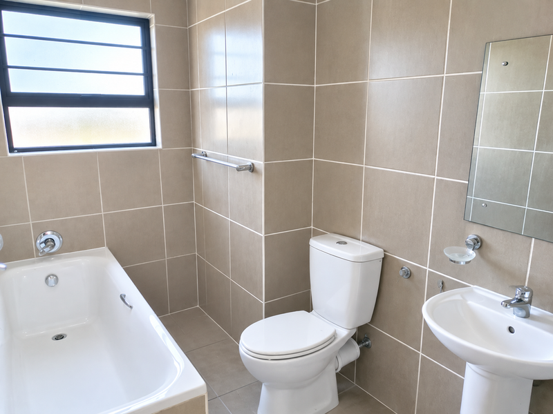 A clean, neutral-toned bathroom featuring a bathtub, pedestal sink, and toilet, finished with floor-to-ceiling wall tiles and a window for natural light.