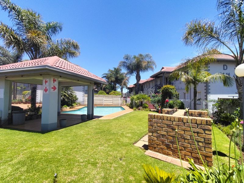Communal outdoor area featuring a swimming pool, covered patio, and a built-in brick braai set within a well-maintained garden.
