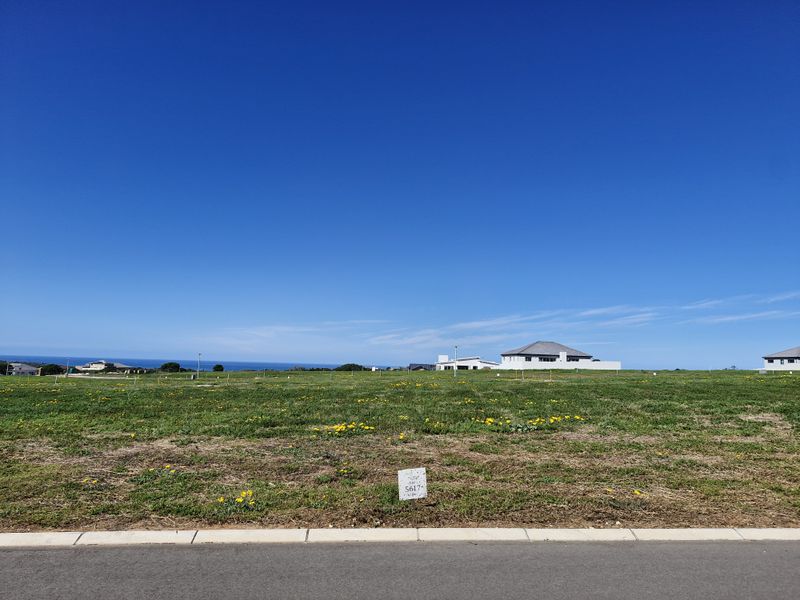 A vacant residential plot featuring a clear view of the surrounding landscape and distant ocean, situated along a paved road with a survey marker.