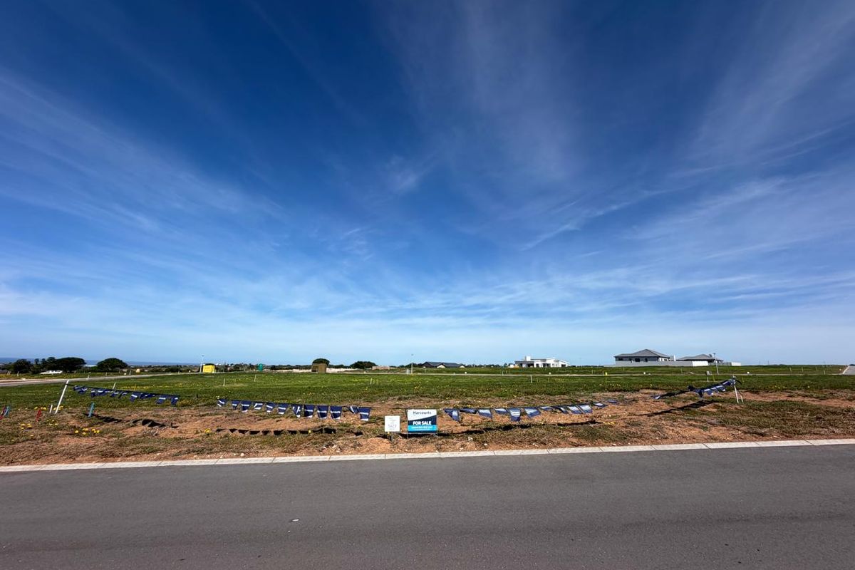 A vacant residential plot of land viewed from the adjacent paved road, featuring open grassed terrain and clear sky.