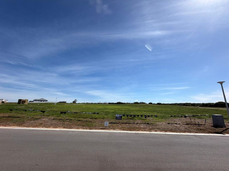 A vacant residential plot of land featuring a level grass surface, accessible from a paved road with existing utility infrastructure.