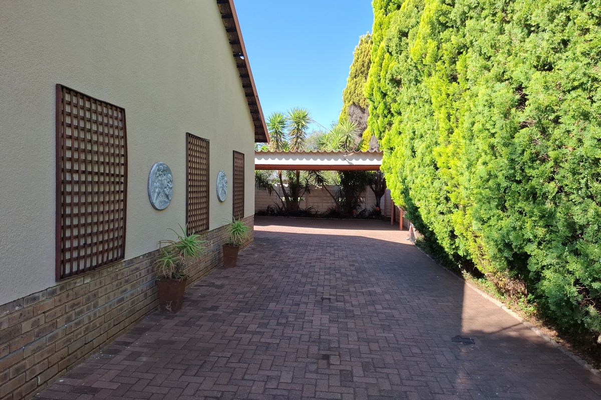 A paved side driveway leading to a rear carport, featuring a brick-based exterior wall with decorative trellis panels and mature conifer hedging.