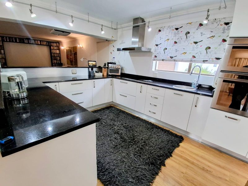 Modern kitchen featuring white cabinetry, black granite countertops, stainless steel appliances, and track lighting, with an open-plan layout connecting to the living area.