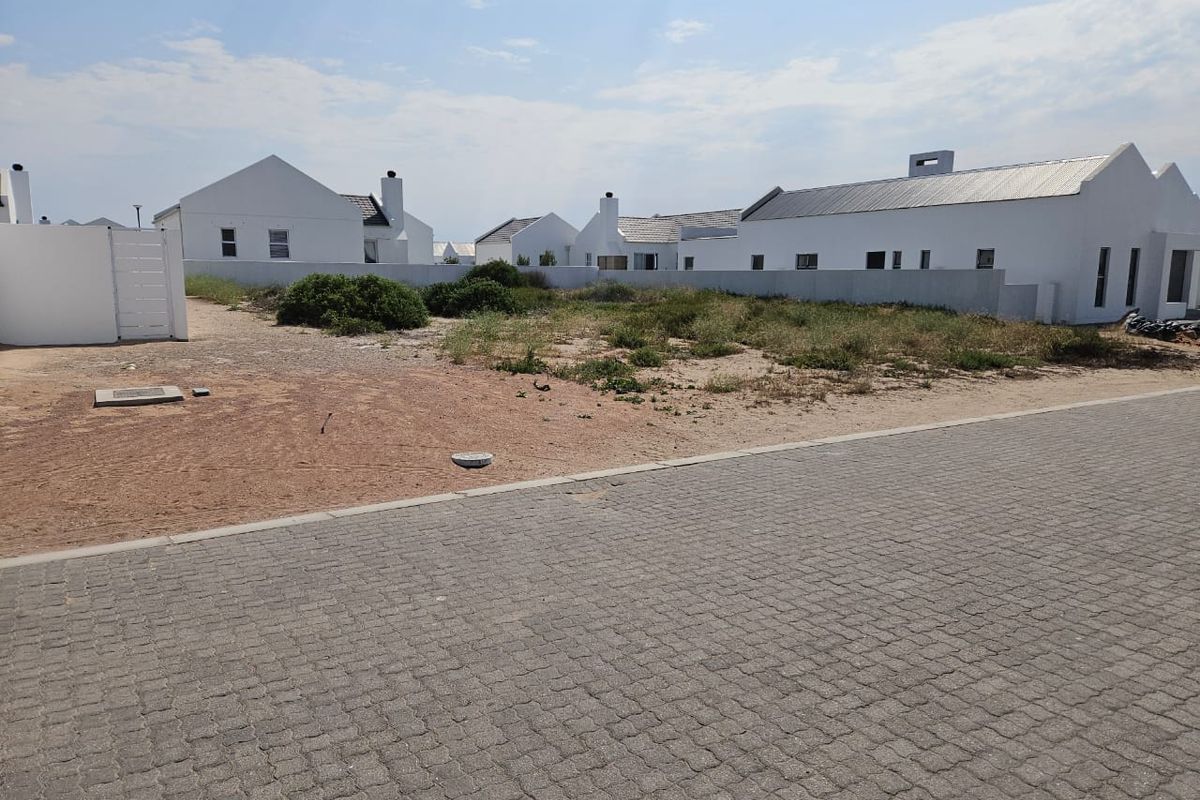 A vacant residential plot situated in a developed estate, featuring paved street access and surrounding boundary walls.
