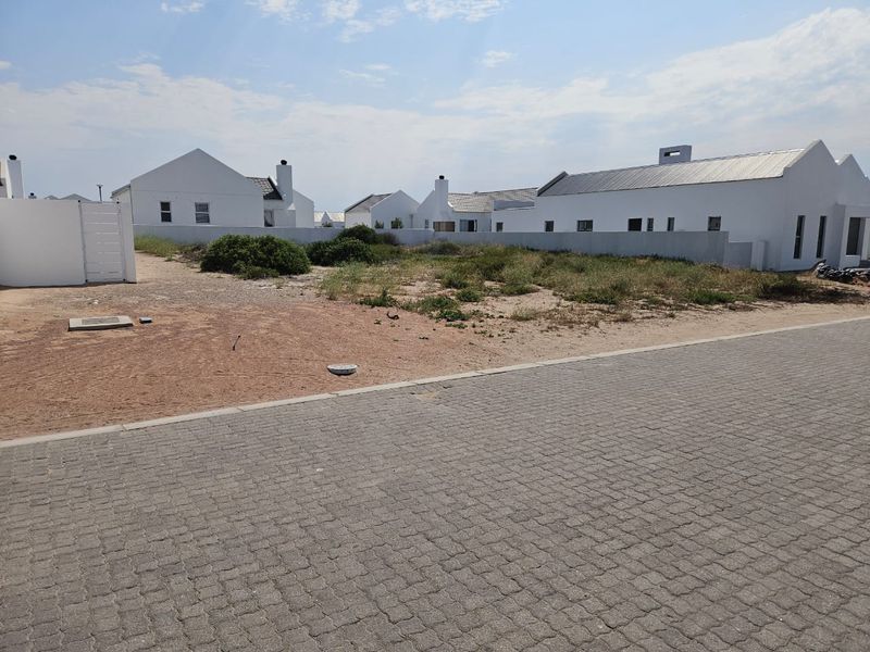 A vacant residential plot situated in a developed estate, featuring paved street access and surrounding boundary walls.