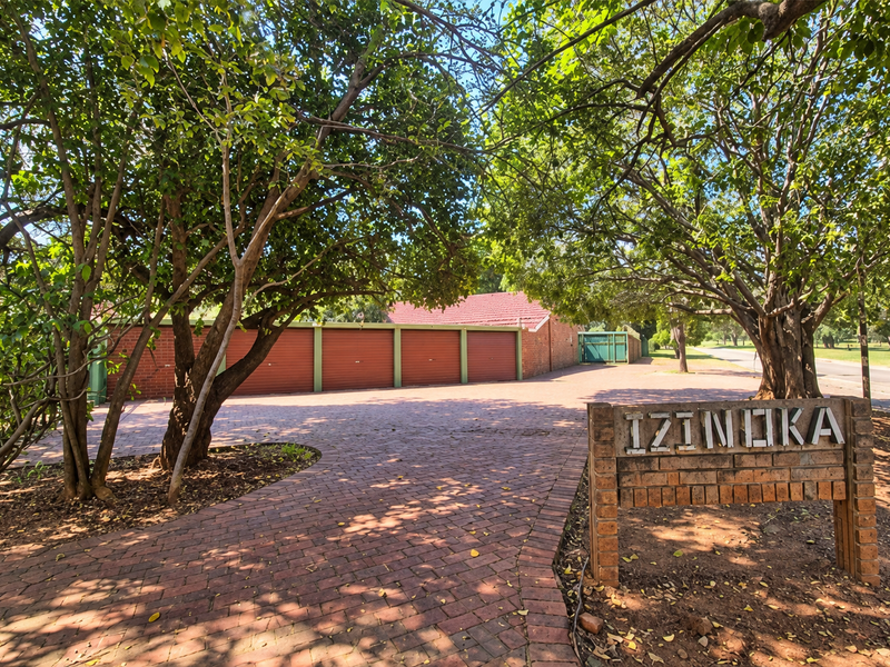 Front entrance view of the apartment complex featuring a brick paved driveway, mature trees, and a row of garages.
