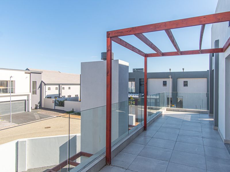 A modern, spacious balcony featuring a wooden pergola, glass balustrade, and tiled flooring, offering an open outdoor space with views of the surrounding estate.