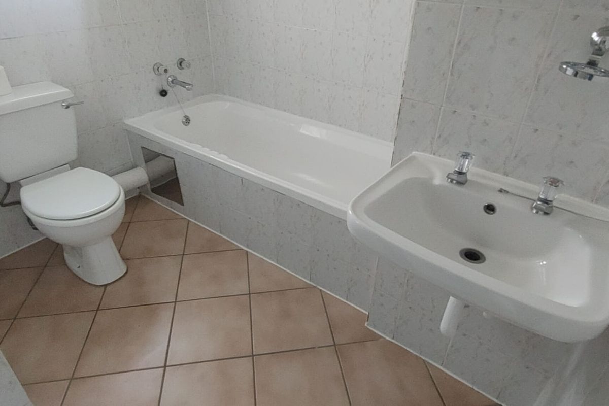A functional bathroom featuring a bathtub, toilet, and wall-mounted basin with tiled finishes, requiring minor maintenance to the bathtub paneling.