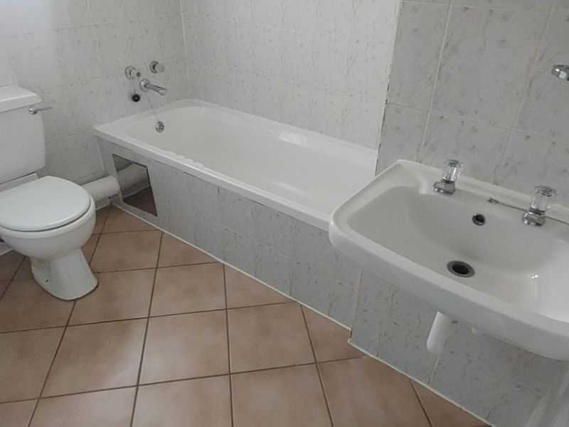 A functional bathroom featuring a bathtub, toilet, and wall-mounted basin with tiled finishes, requiring minor maintenance to the bathtub paneling.