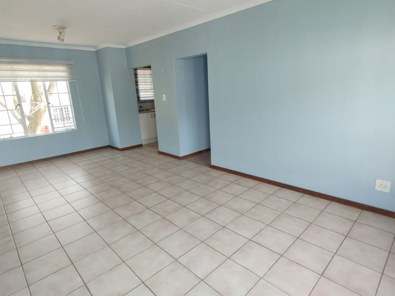 A spacious, empty living room featuring neutral tiled flooring, light blue walls, and a window with a roller blind, offering a clean, blank canvas.
