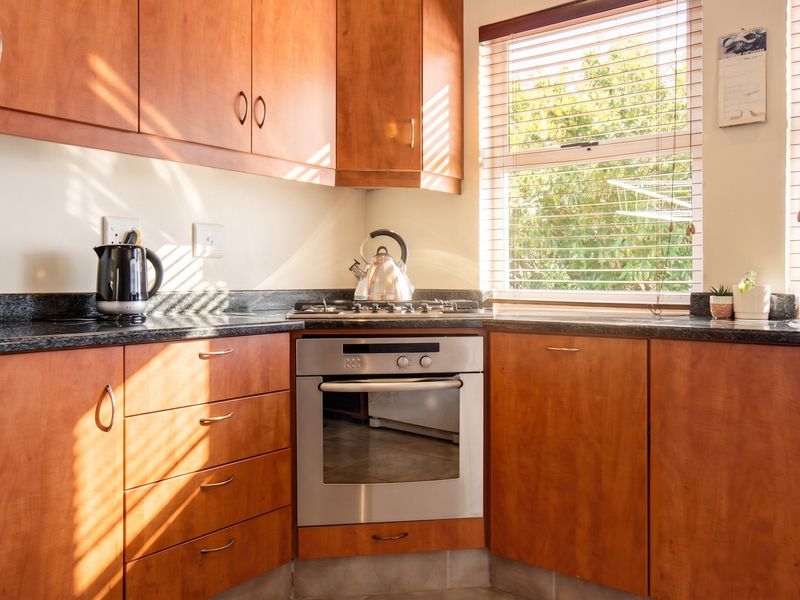 A functional kitchen featuring warm-toned wooden cabinetry, granite countertops, and a built-in oven with a gas hob, illuminated by natural light through window blinds.