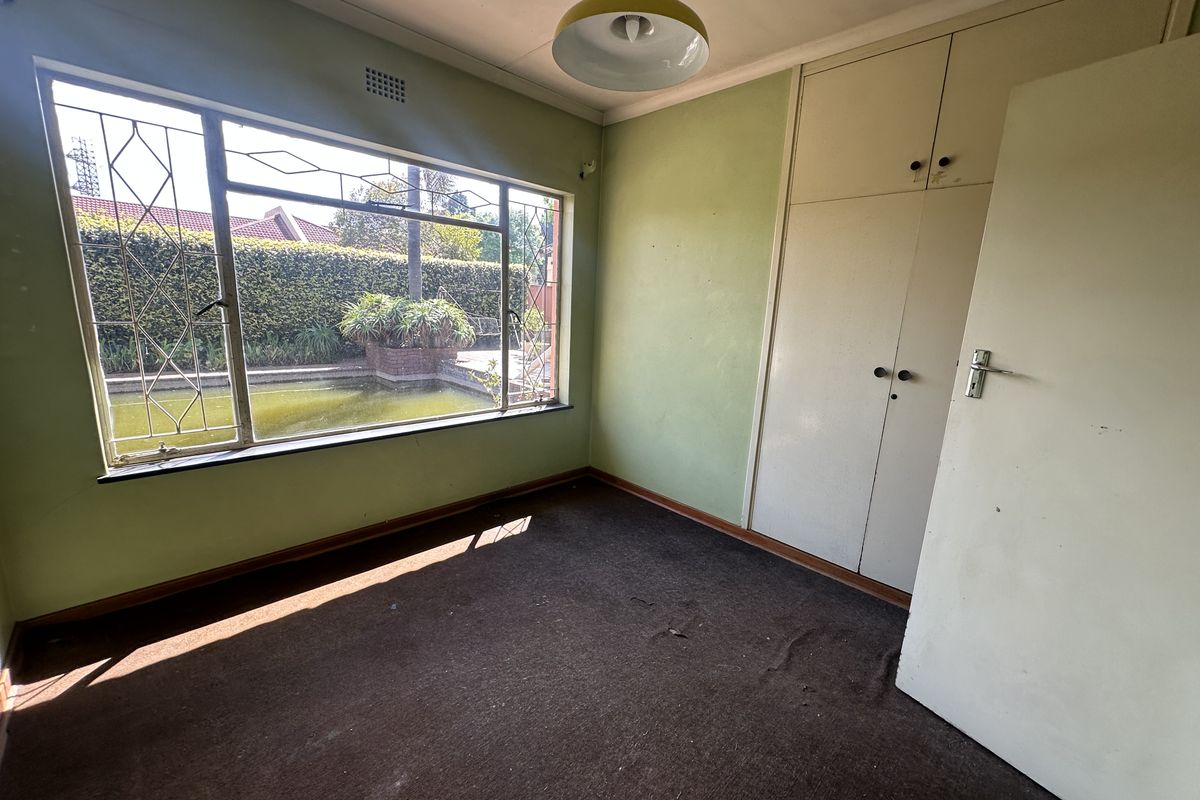 An empty bedroom featuring built-in cupboards, carpet flooring, and a large window with security bars overlooking the garden area.