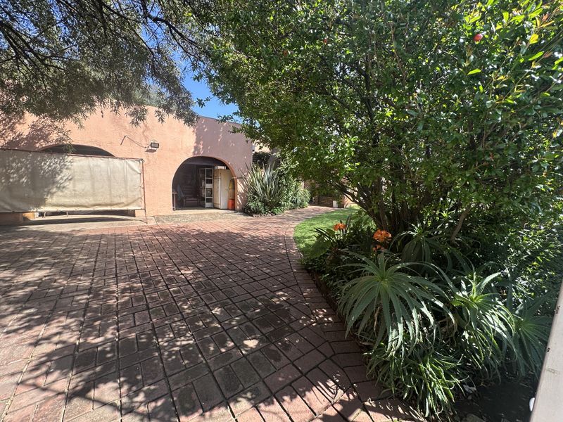 A rear exterior view featuring a brick-paved patio area leading to a covered arched space, surrounded by established garden landscaping.