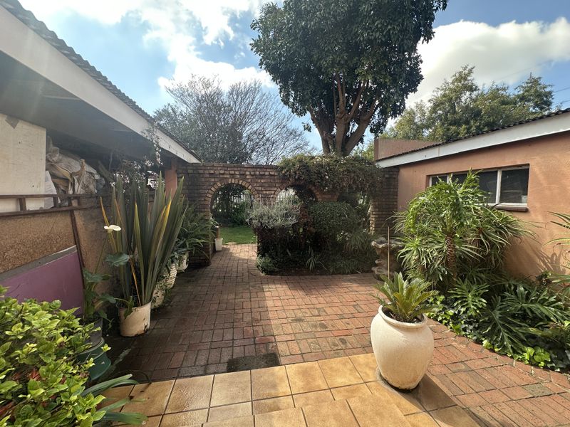 A paved outdoor patio area featuring a decorative brick archway, established garden beds, and tiled steps leading into the courtyard space.