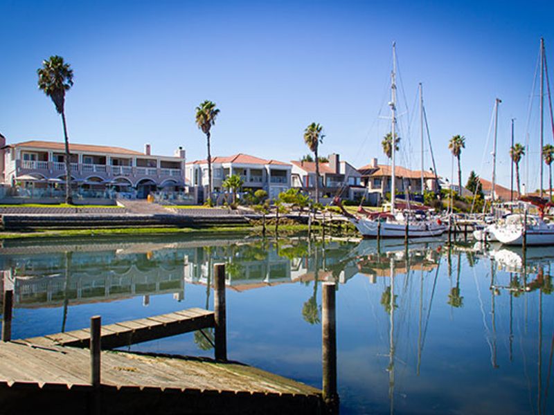 A scenic view of a residential waterfront estate featuring a private jetty, calm marina waters, and docked sailboats under a clear blue sky.