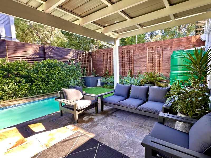 A covered outdoor patio area featuring paved flooring, overlooking a private swimming pool and enclosed by timber fencing with a water tank.