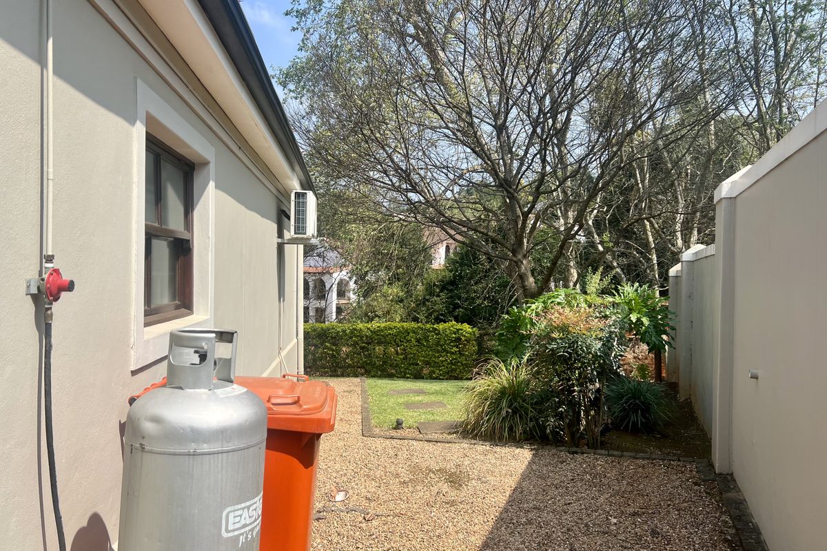 A side exterior view of the property featuring a gravel pathway, small lawn area, boundary wall, and an external air conditioning unit.