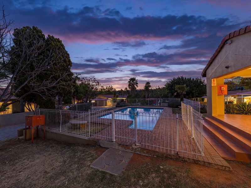 A twilight view of a residential swimming pool area featuring paved surrounds, safety fencing, and an adjacent covered patio space.