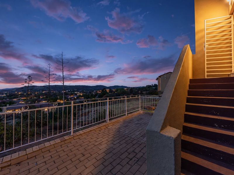 An elevated outdoor balcony featuring paved flooring, a metal balustrade, and an external staircase, offering scenic sunset views over the surrounding neighborhood.
