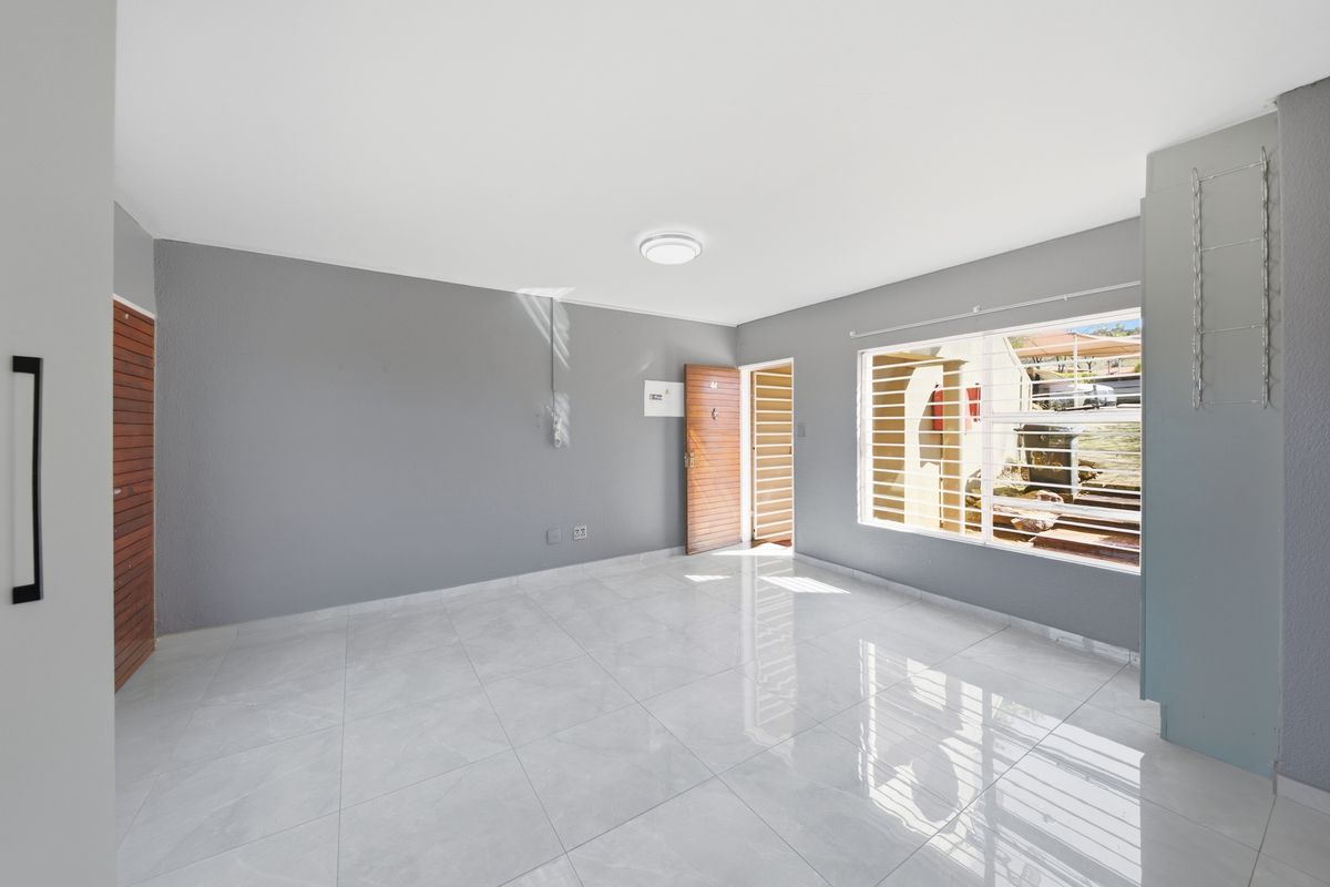A bright, empty living area featuring neutral grey walls, polished tiled flooring, and natural light through a window with security bars.